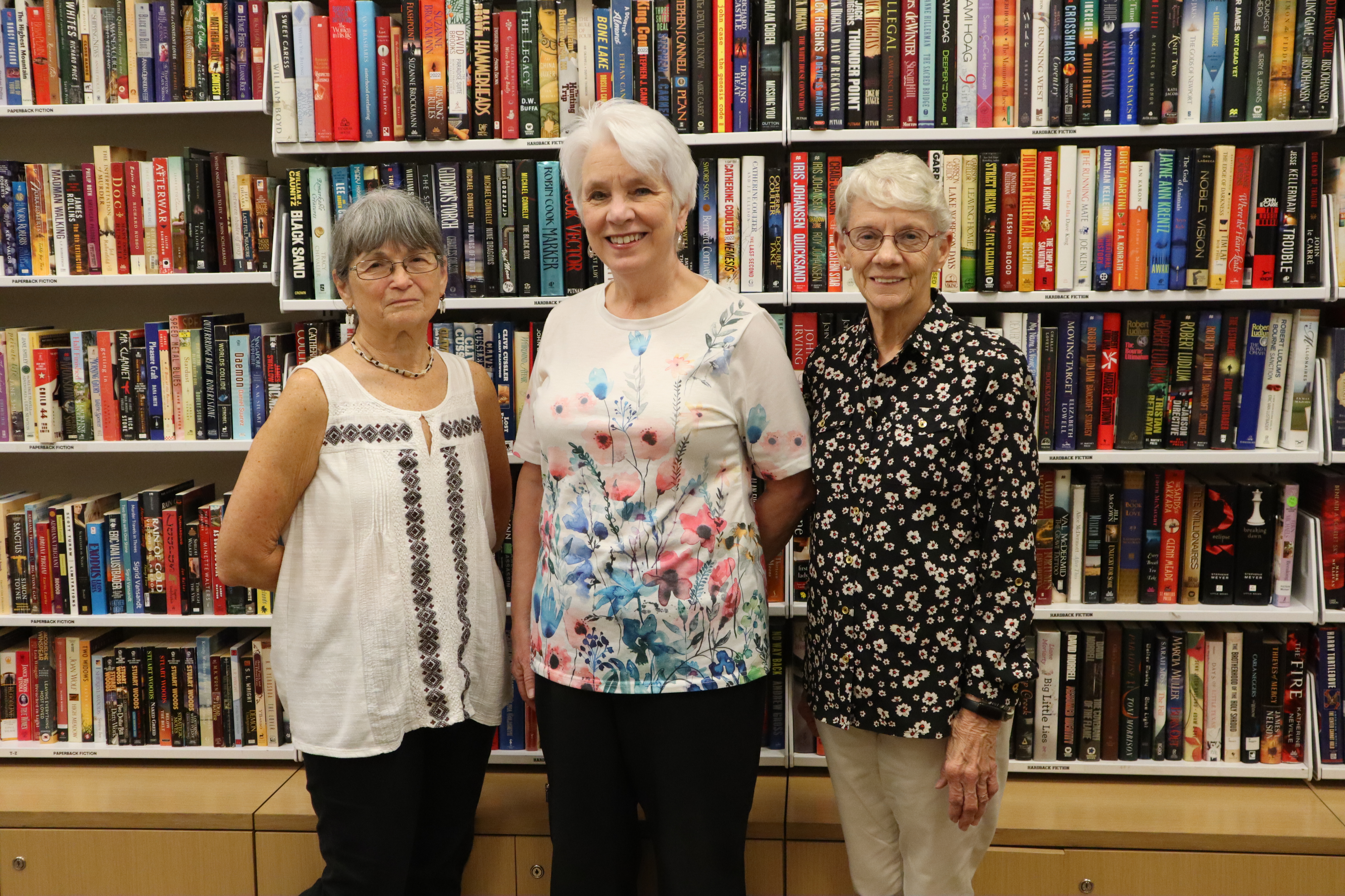 Three McAllen Public Library volunteers stand smiling in front of shelves filled with fiction books, posing together inside the library.